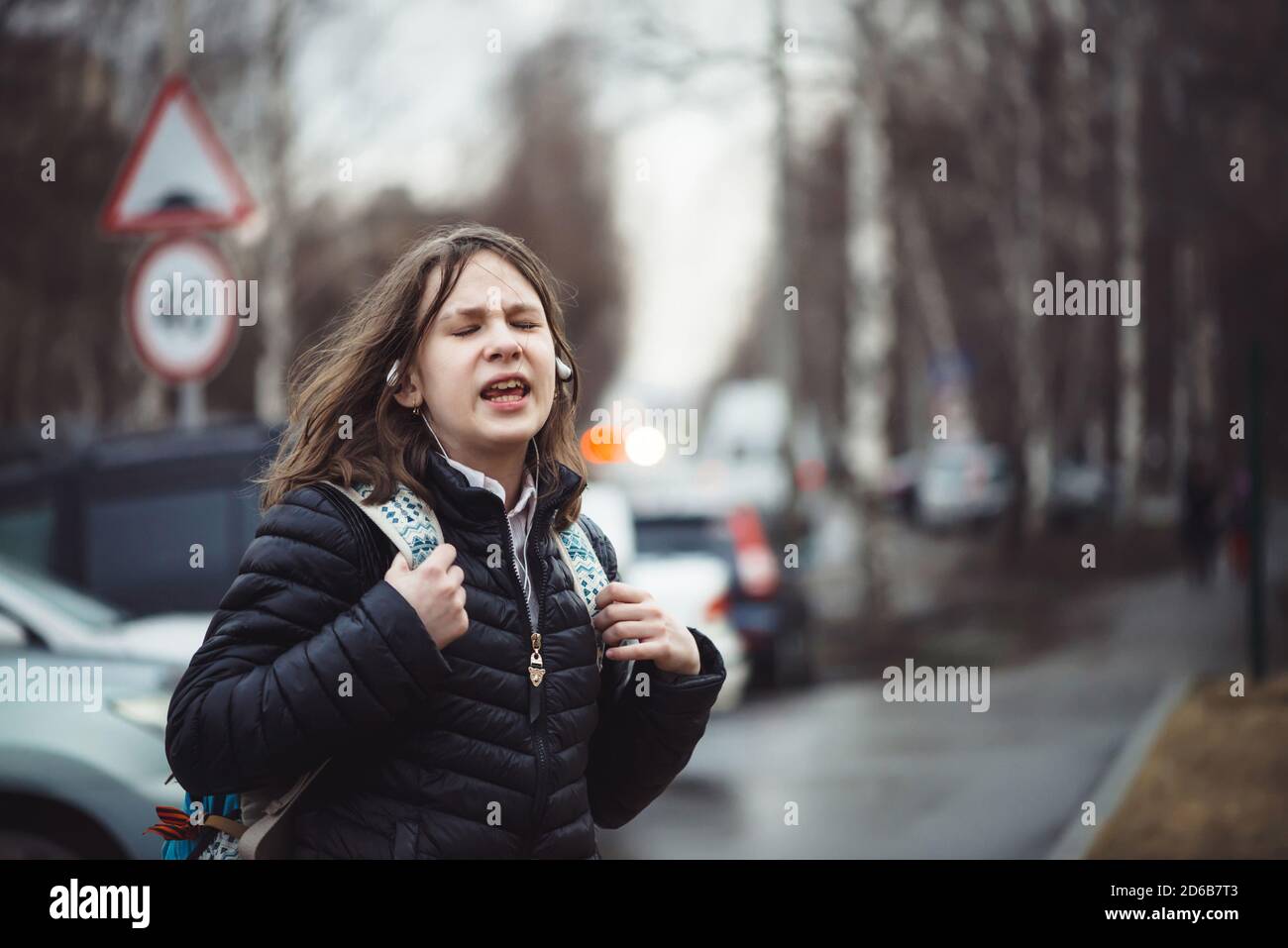 Emotional explosion of capricious teen. Crazy schoolgirl with backpack in depressed state in city in rainy weather. Emotion surge swept young girl wit Stock Photo