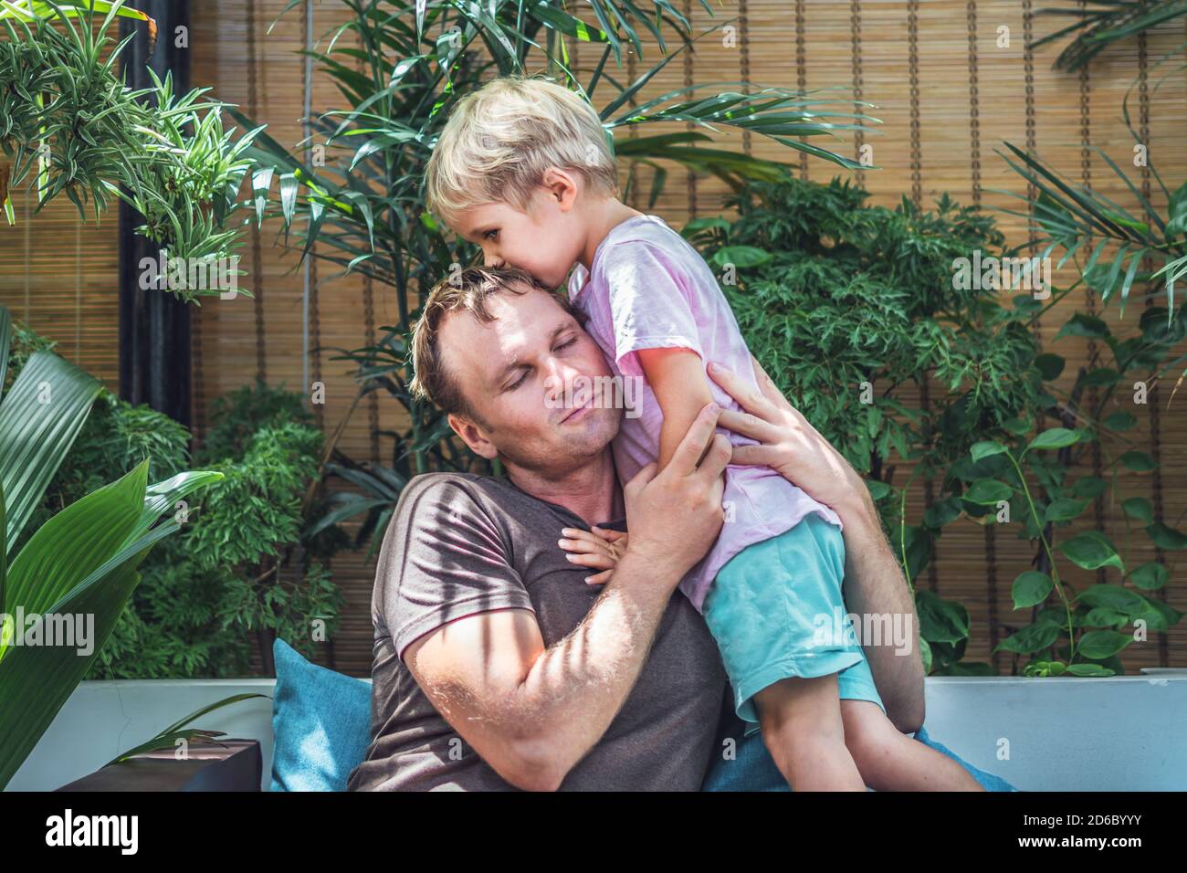 Father gently hugs son, eyes closed with pleasure, blond little child boy kiss daddy head top, face to face, bright sun light green leaves back. Happy Stock Photo