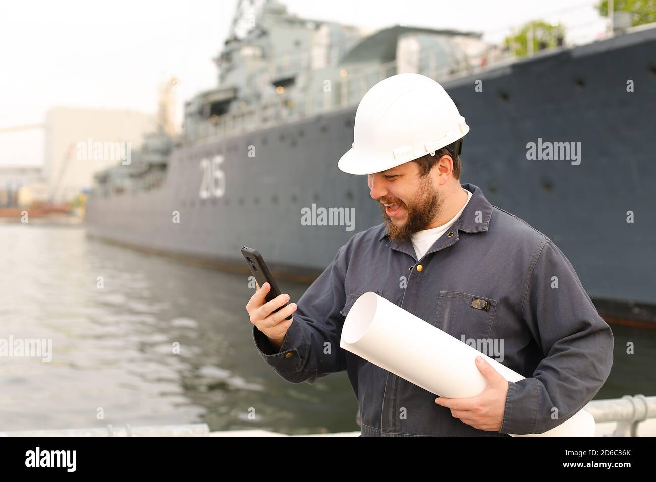 Marine happy engineer talking by VHF walkie talkie, holding papers near vessel in background. Stock Photo