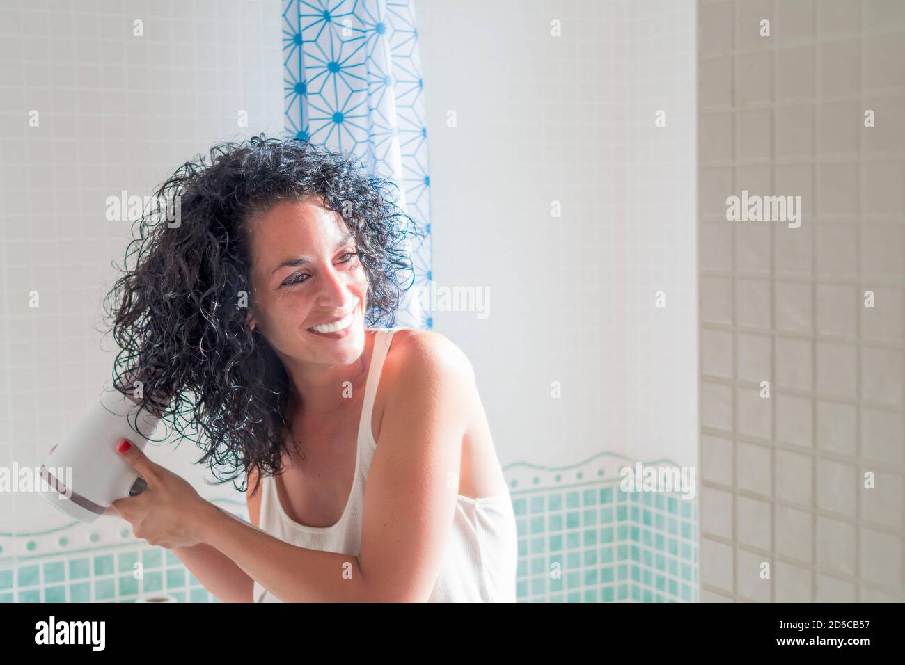 young woman smiling in front of the mirror curling her hair with a hairdryer with a special nozzle to curl her hair. dryer with diffuser. care and bea Stock Photo