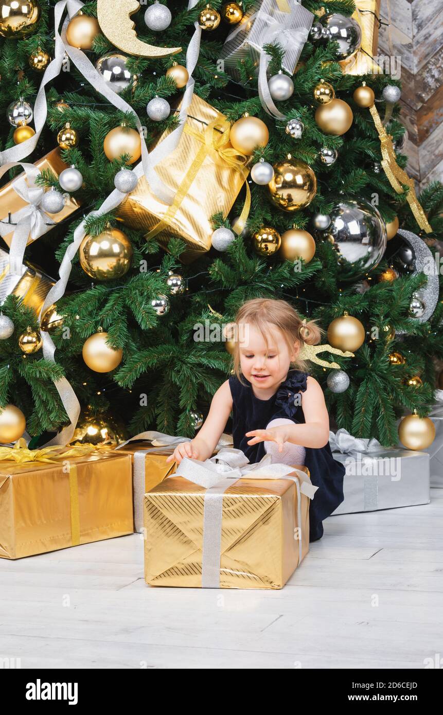 Portrait of elementary age girl wearing blue dress sitting beside christmas tree unpacking big wrapped gift box curious about a present on boxing day Stock Photo