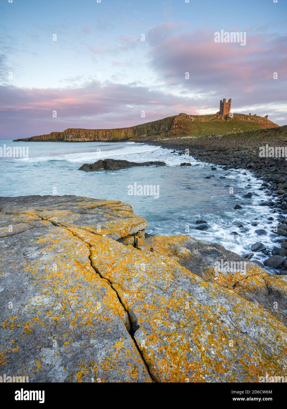 Dunstanburgh Castle glows in late evening light at sunset, with the weathered, lichen covered rocks of Embleton Bay visible in the foreground. Stock Photo