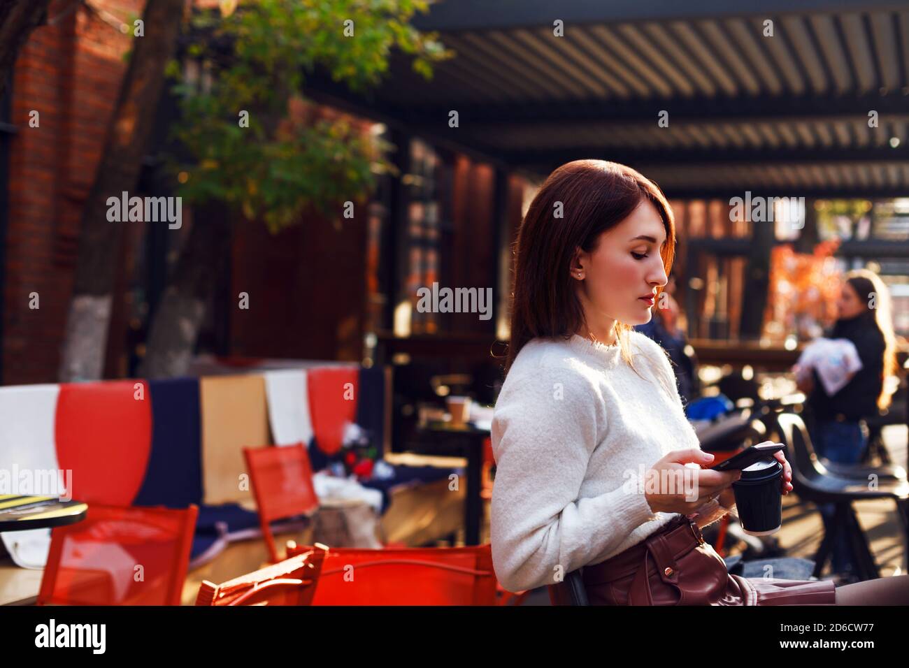 Woman sits at outdoor cafe terrace with takeaway coffee and swipes something on her smartphone. Stock Photo