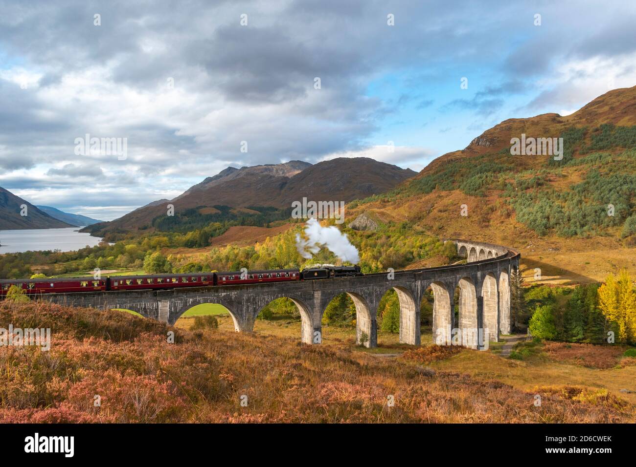 FORT WILLIAM SCOTLAND THE JACOBITE STEAM TRAIN CROSSING THE GLENFINNAN VIADUCT WITH AUTUMN COLOURS IN THE SCENERY AND LOCH SHIEL IN THE DISTANCE Stock Photo