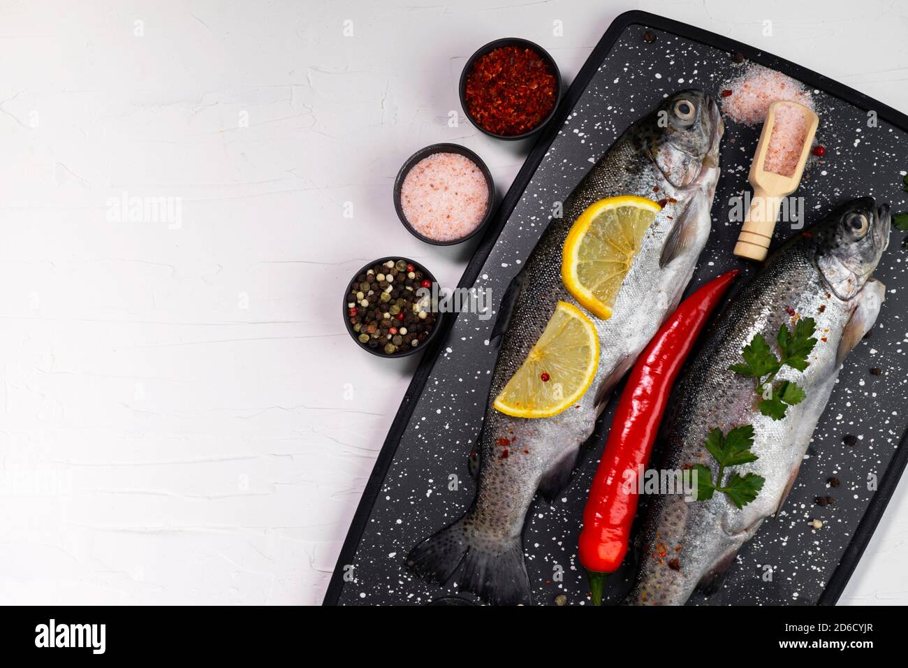 Fresh rainbow trouts on black board on table with salt, pepper, lemon and chili pepper ingredients, flatlay, copy space Stock Photo