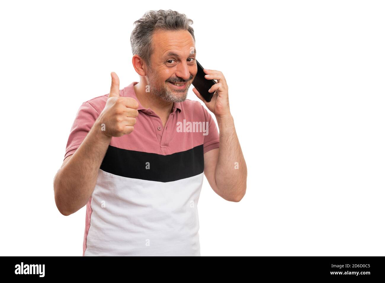 Friendly adult man smiling as talking on mobile phone making thumb up like gesture wearing fashionable summer attire tshirt isolated on white backgrou Stock Photo