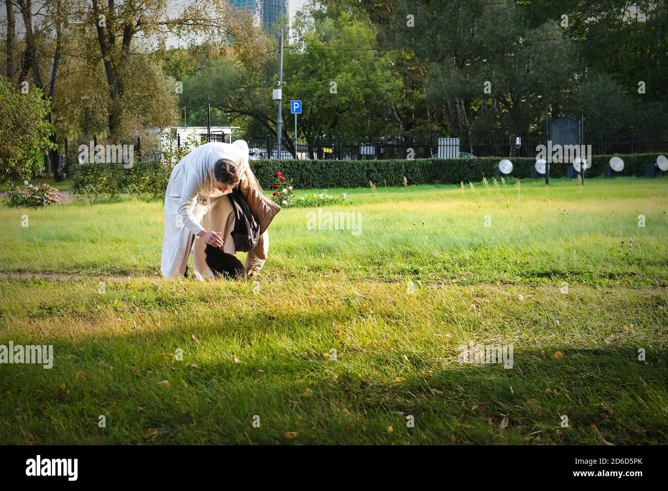 Caring greybeard cleric wearing white priestly garb stroking stray black cat after divine service in church park.  Stock Photo