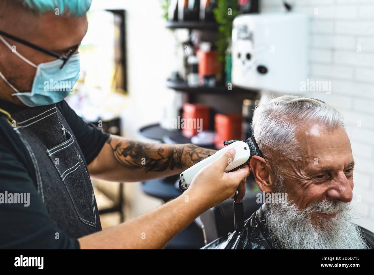 Male hairdresser cutting hair to hipster senior client while wearing face surgical mask - Young hairstylist working in barbershop during corona virus Stock Photo