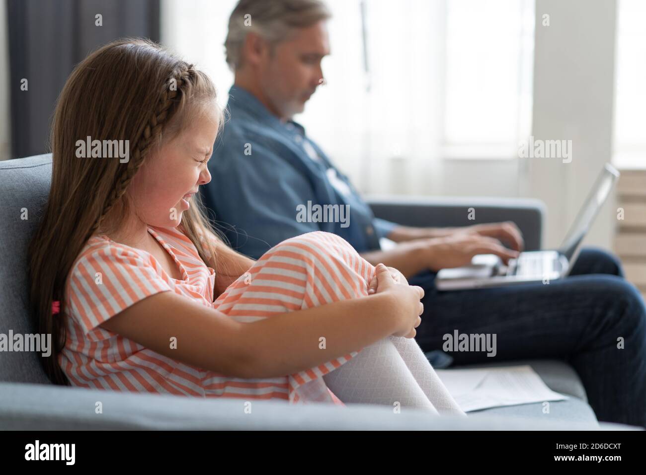 No Time For Child. Grey-haired dad busy with laptop, working online at home, sad bored offended daughter sitting near by Stock Photo