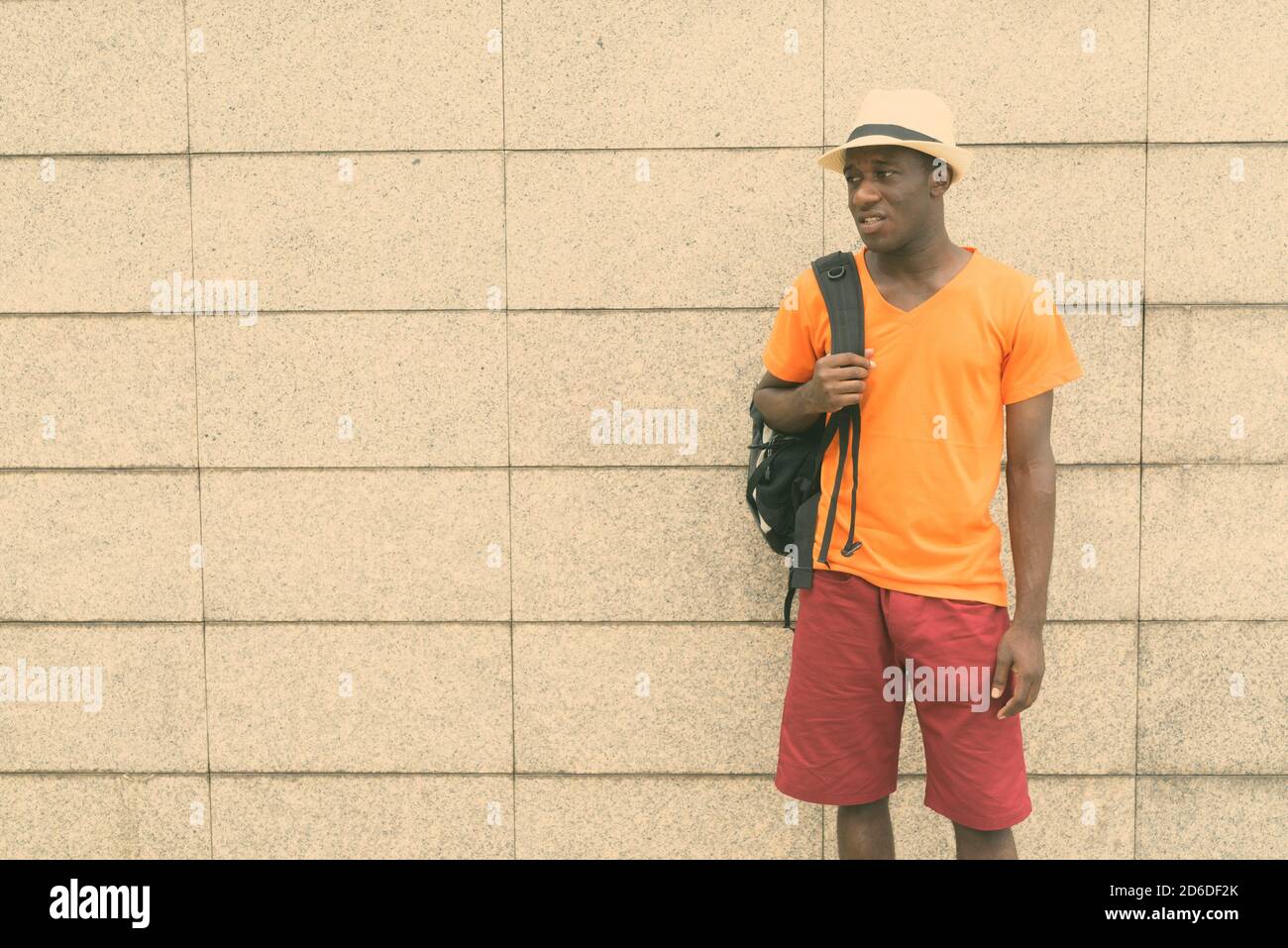 Young black African tourist man standing and thinking while holding backpack against concrete block wall Stock Photo