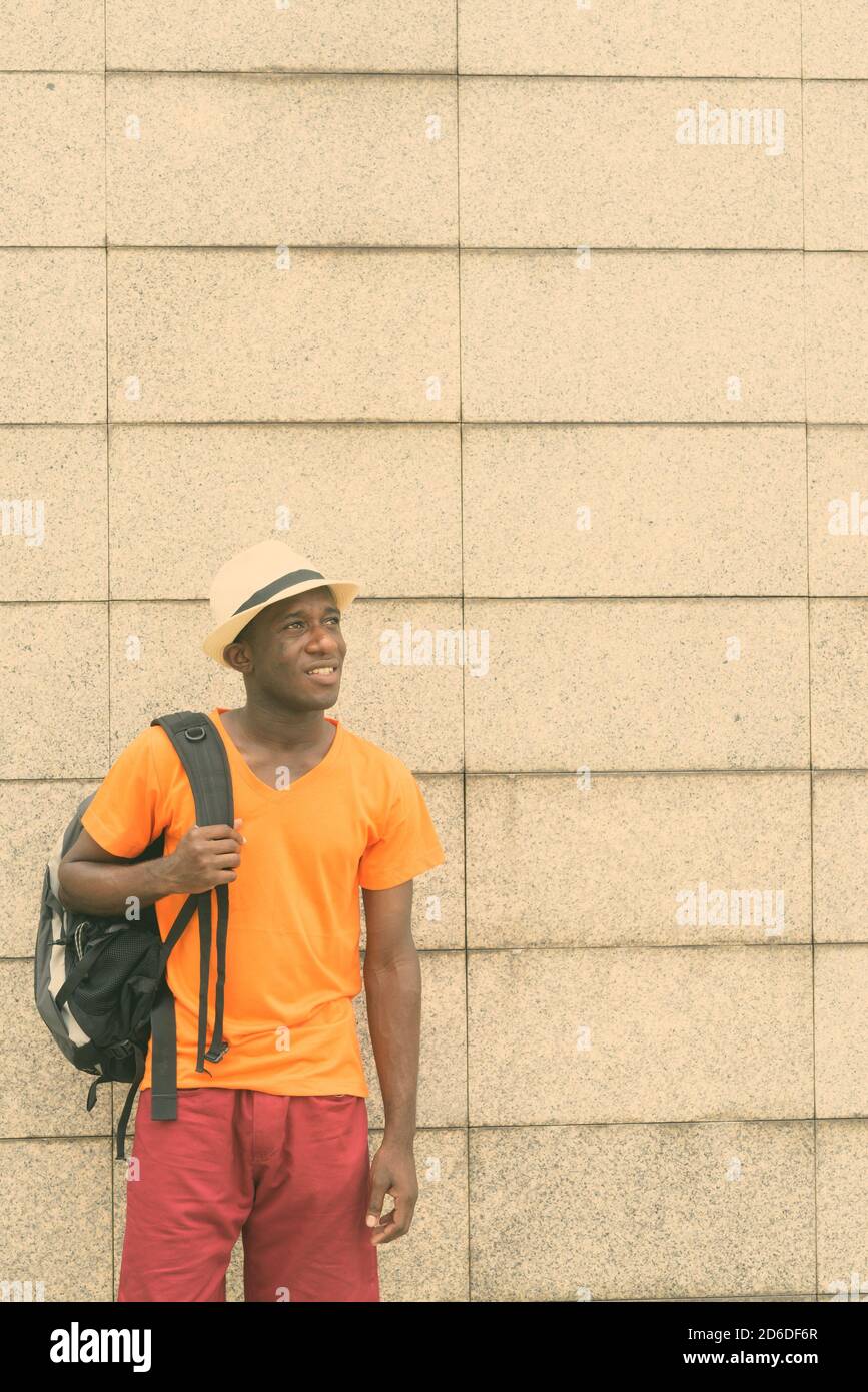 Young happy black African tourist man smiling while thinking and holding backpack against concrete block wall Stock Photo