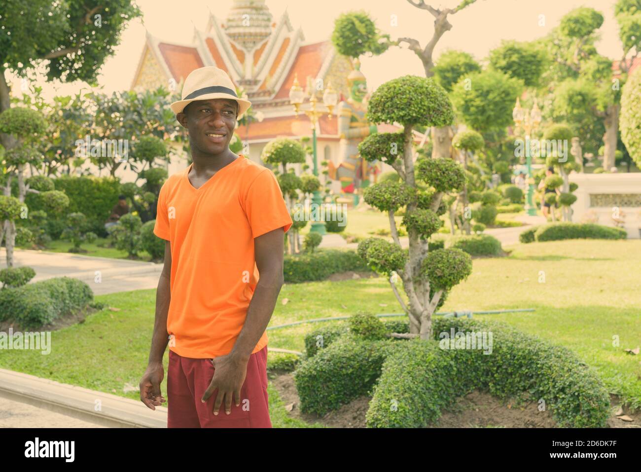 Young happy black African tourist man smiling while thinking against view of beautiful garden and Wat Arun temple in Bangkok Thailand Stock Photo