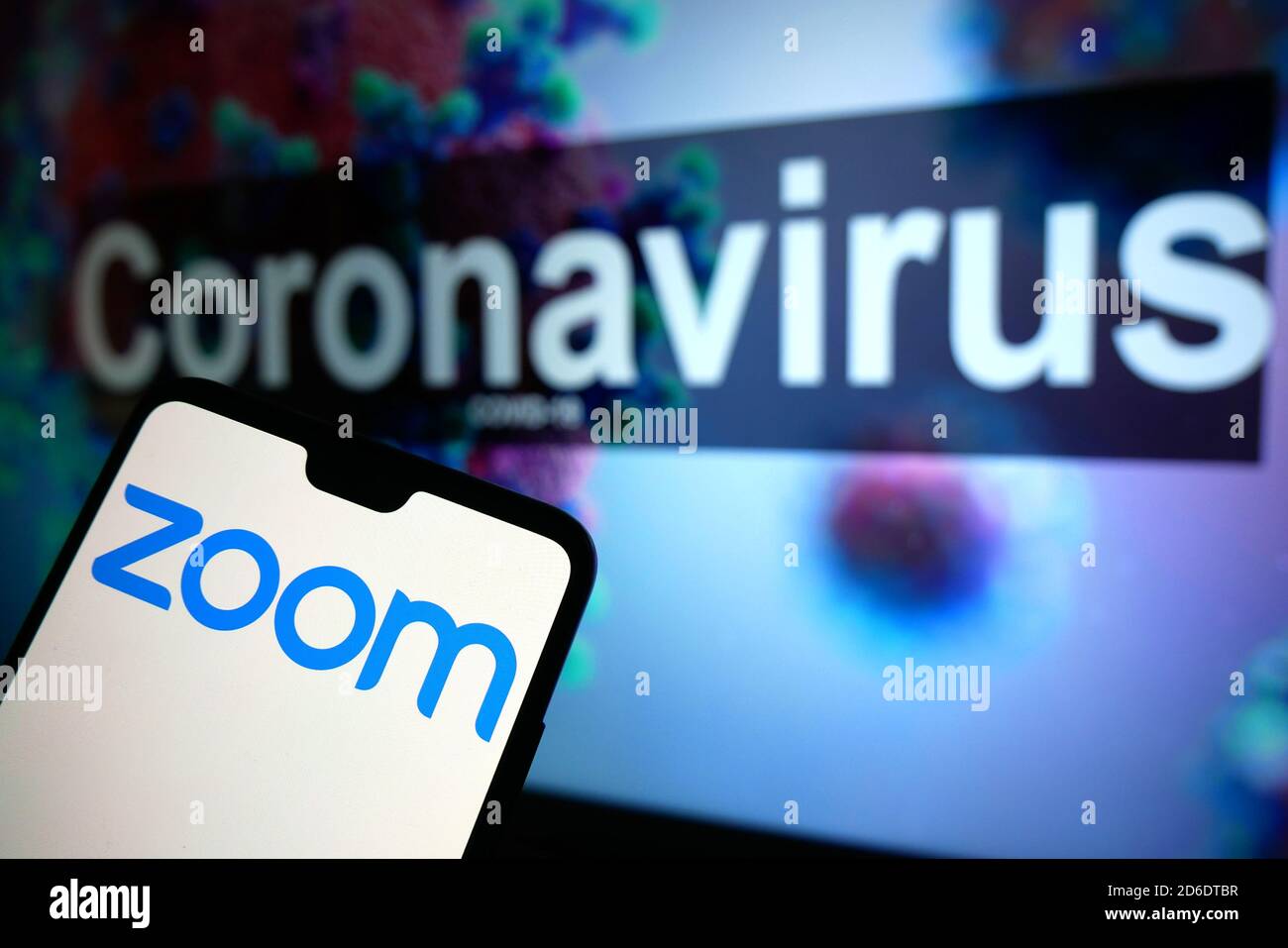 The Zoom logo seen displayed on a mobile phone with an illustrative model of the Coronavirus displayed on a monitor in the background. Stock Photo