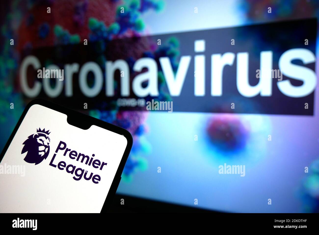 The Premier League logo seen displayed on a mobile phone with an illustrative model of the Coronavirus displayed on a monitor in the background. Stock Photo