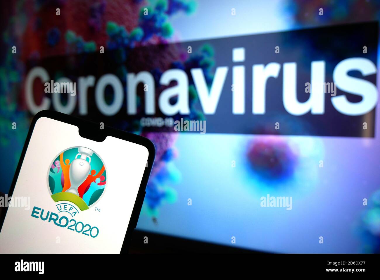 The UEFA Euro 2020 logo seen displayed on a mobile phone with an illustrative model of the Coronavirus displayed on a monitor in the background. Stock Photo