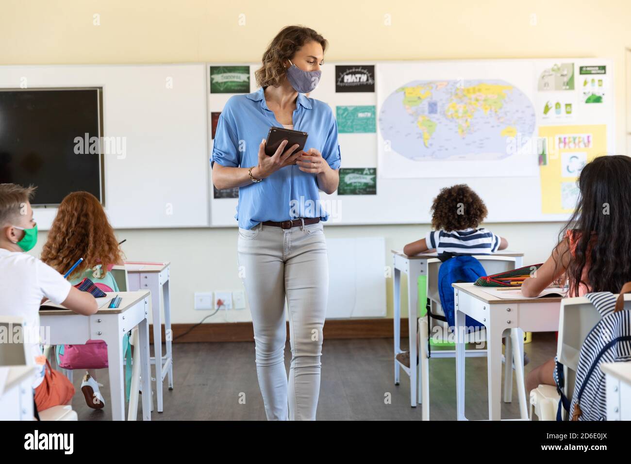 Female teacher wearing face mask holding digital tablet teaching to students in class Stock Photo