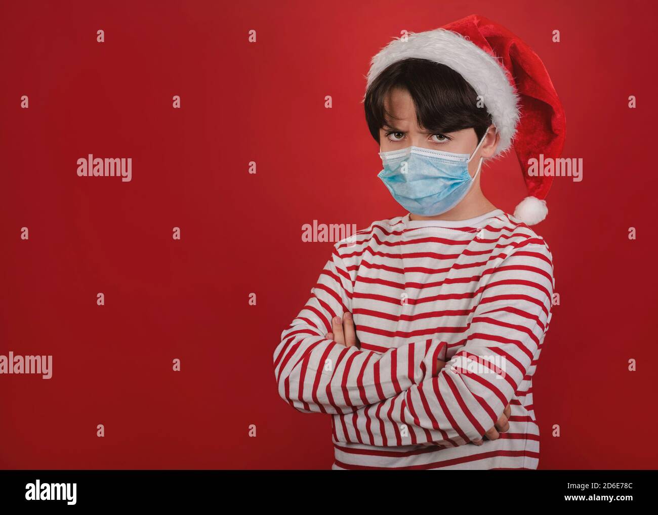 angry kid with medical mask wearing Santa Claus Hat over red background Stock Photo