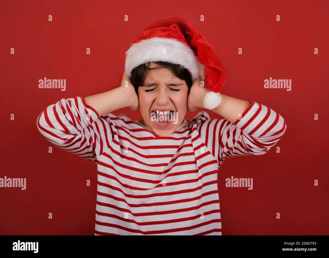 angry kid wearing Santa Claus hat with his hands on his head over red background Stock Photo