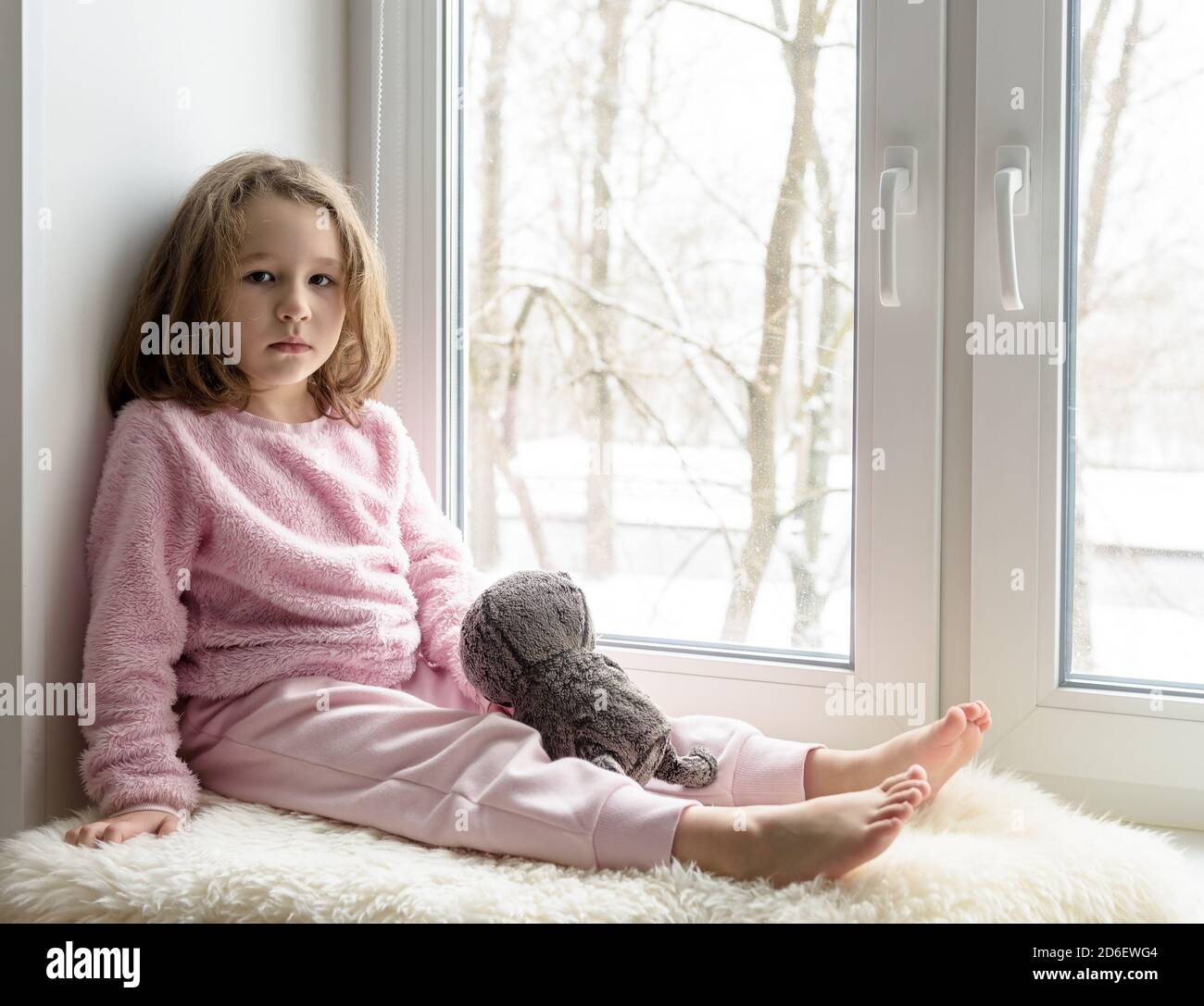 Kid sits on windowsill at home and looks at camera, portrait of pretty little girl on fur rug on room sill in winter. Adorable pensive child in pink, Stock Photo