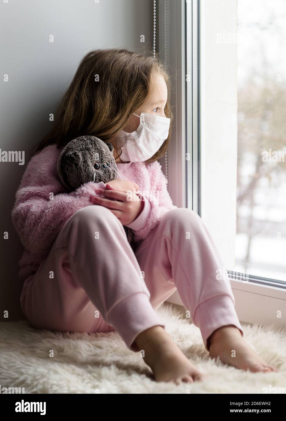 COVID-19 coronavirus concept, sad kid in medical mask looks out window in house. Pensive child sits on windowsill at home in winter. Little girl with Stock Photo