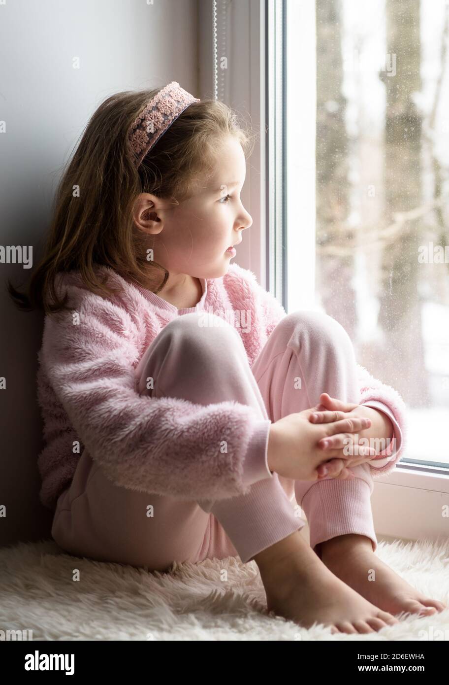 Kid sitting on windowsill at home looks out window to snow, portrait of pretty little girl on fur rug on room sill in winter. Adorable pensive child i Stock Photo