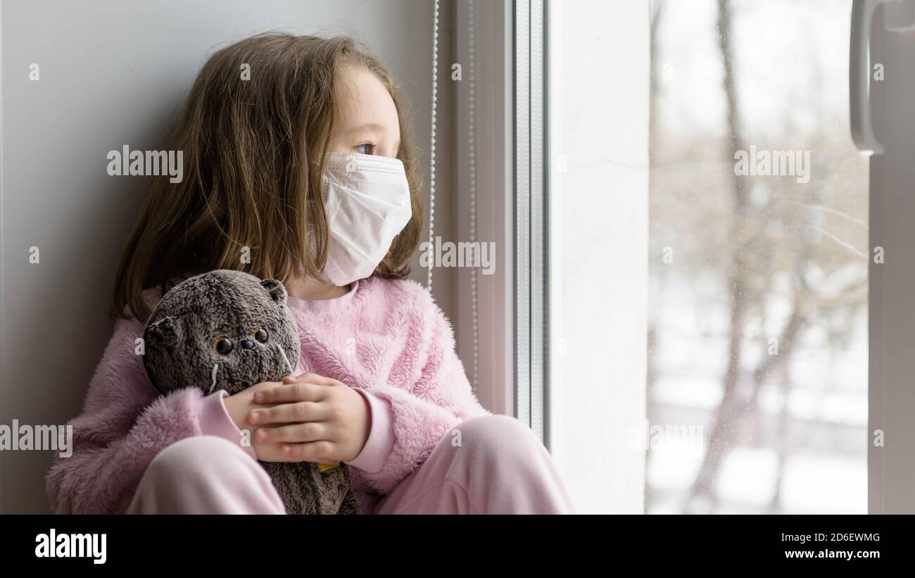COVID-19 coronavirus concept, sad kid in medical mask looks out window. Pensive child sits on windowsill at home in autumn or winter. Little girl with Stock Photo