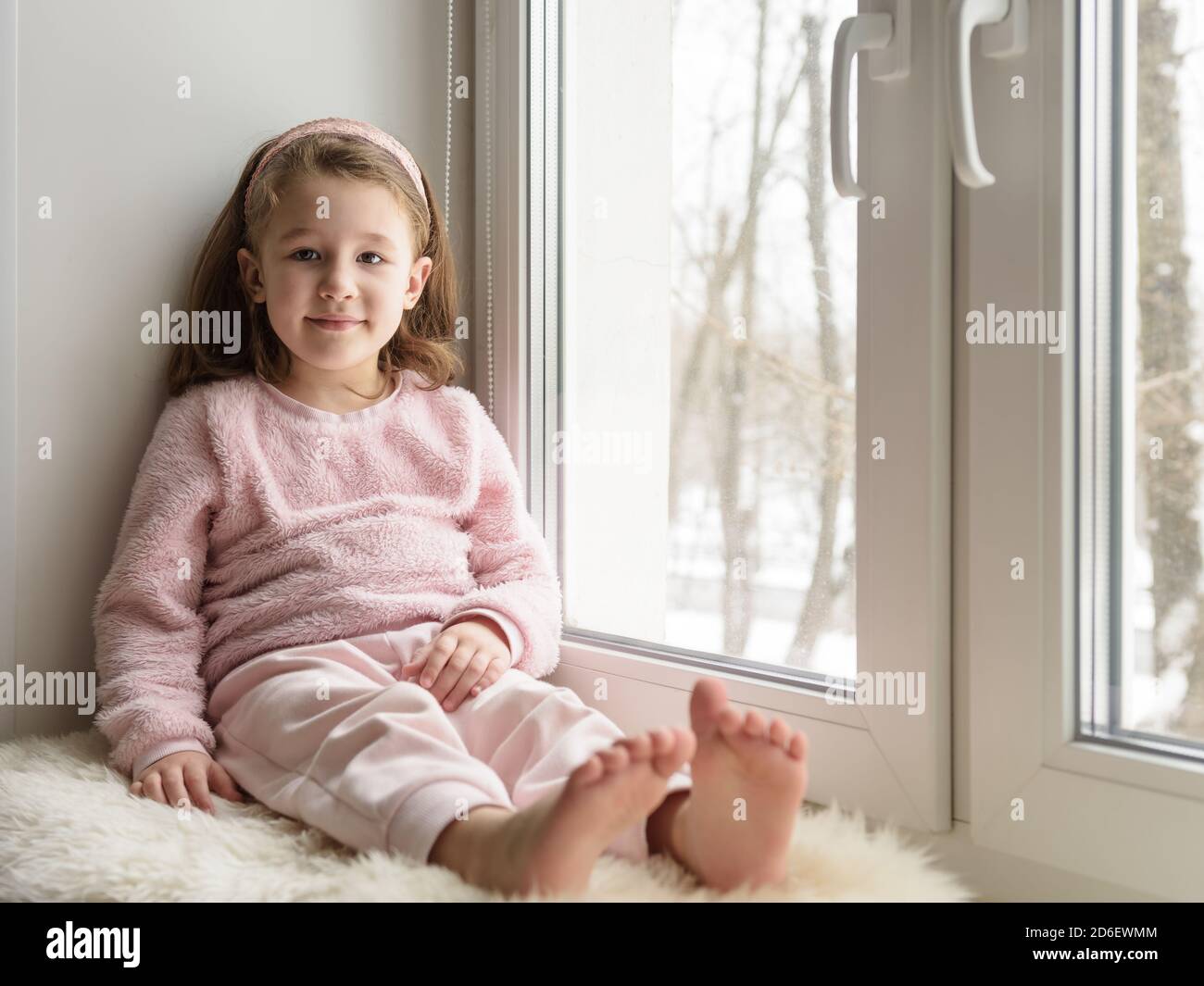 Kid sitting by window looks at camera indoor, portrait of pretty little girl on fur rug on room sill in winter. Adorable smiling child in pink is on w Stock Photo