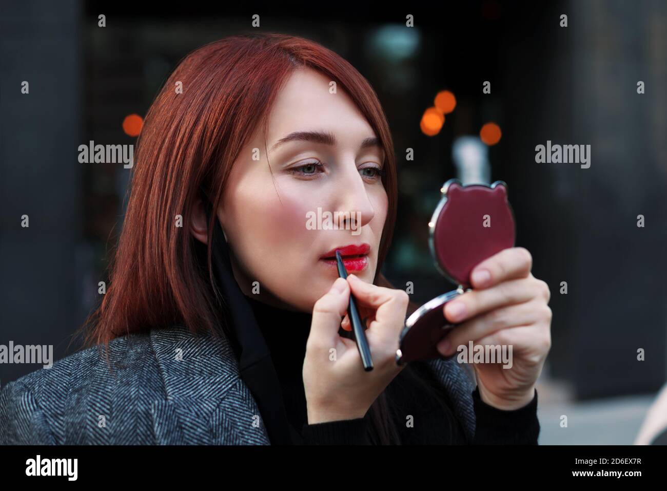 Pretty redhead woman looks at pocket mirror and applys red lipstic before business meeting. Dating after work concept. Stock Photo