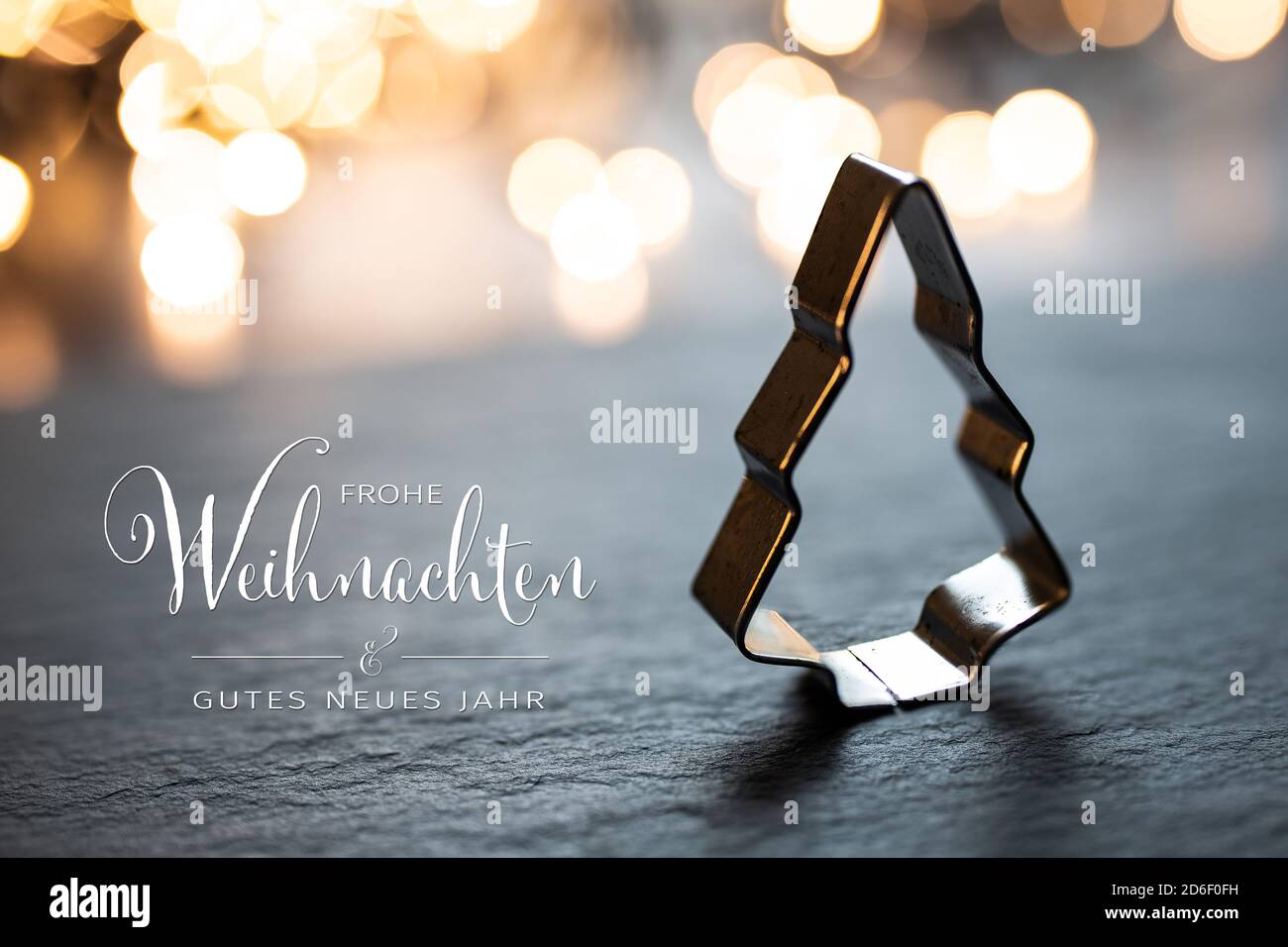 Christmas Tree Cookie cutter in front of christmas lights. Text 'Merry Christmas & Happy New Year' in German 'Frohe Weihnachten & Gutes Neues Jahr' to Stock Photo