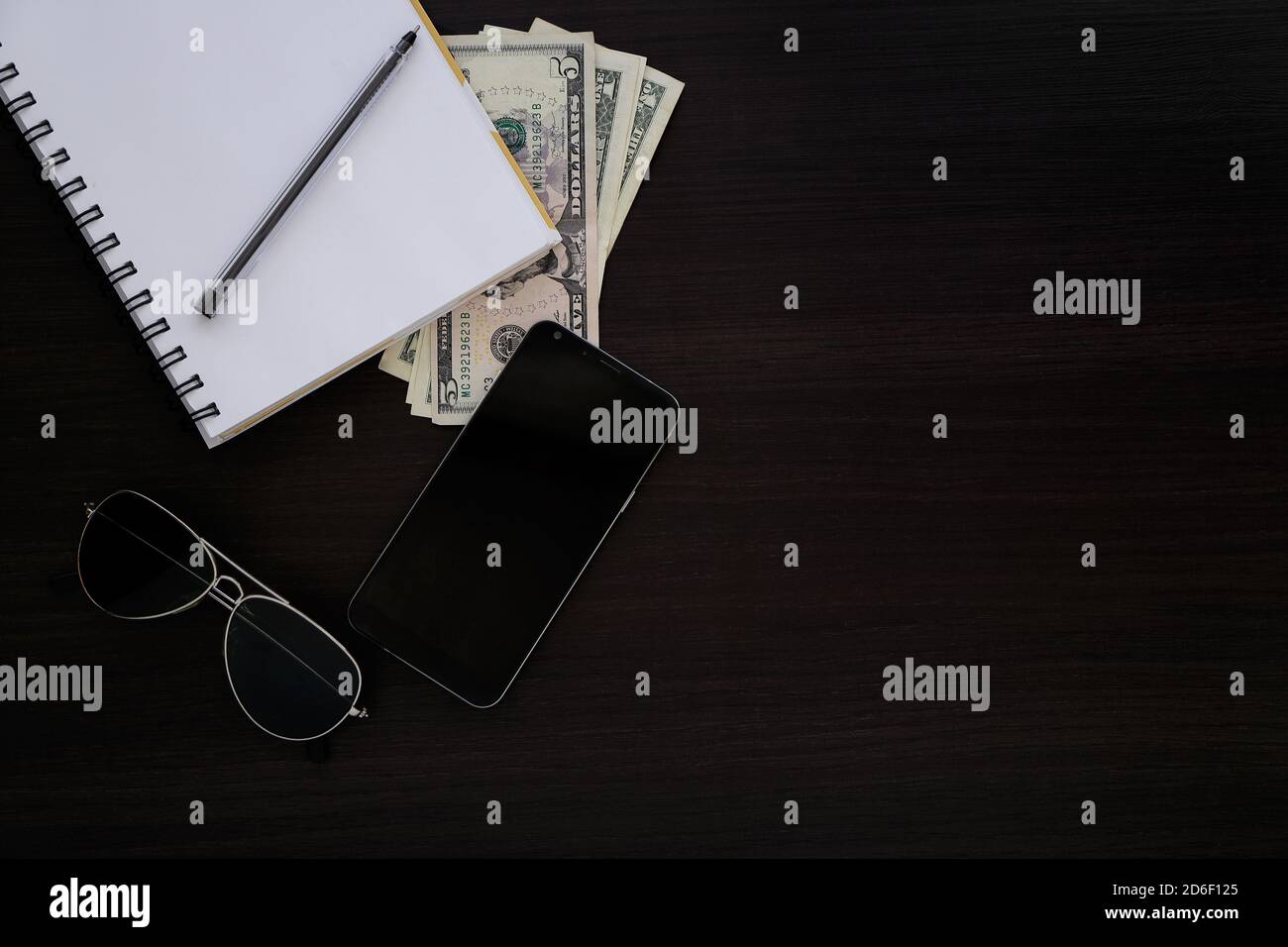 Dark wooden desk with objects. Top view of a notebook, money, cell, phone, pen and sunglasses. Stock Photo