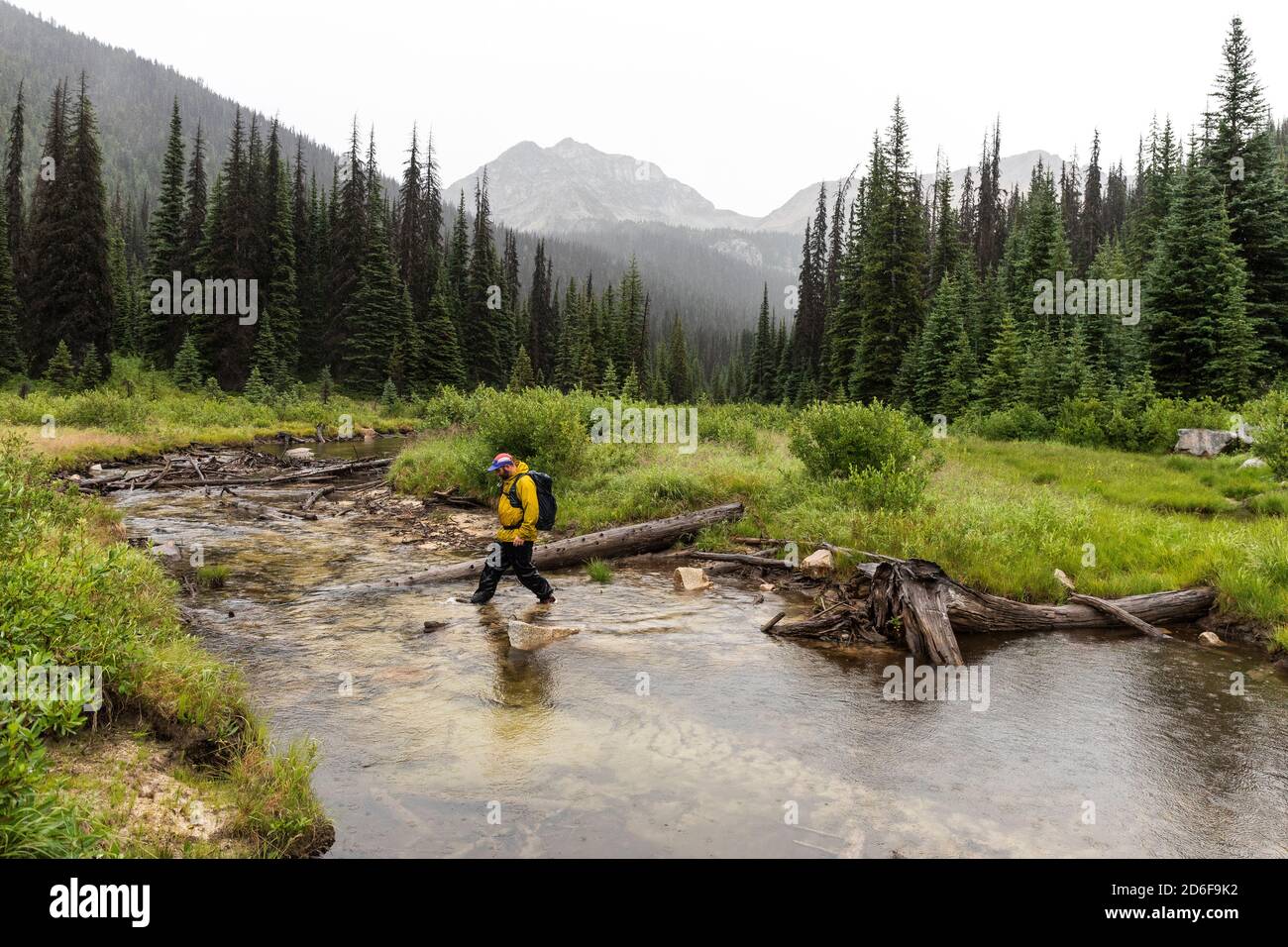 Side view of man hiking in mountains and crossing river on overcast day in British Columbia Stock Photo