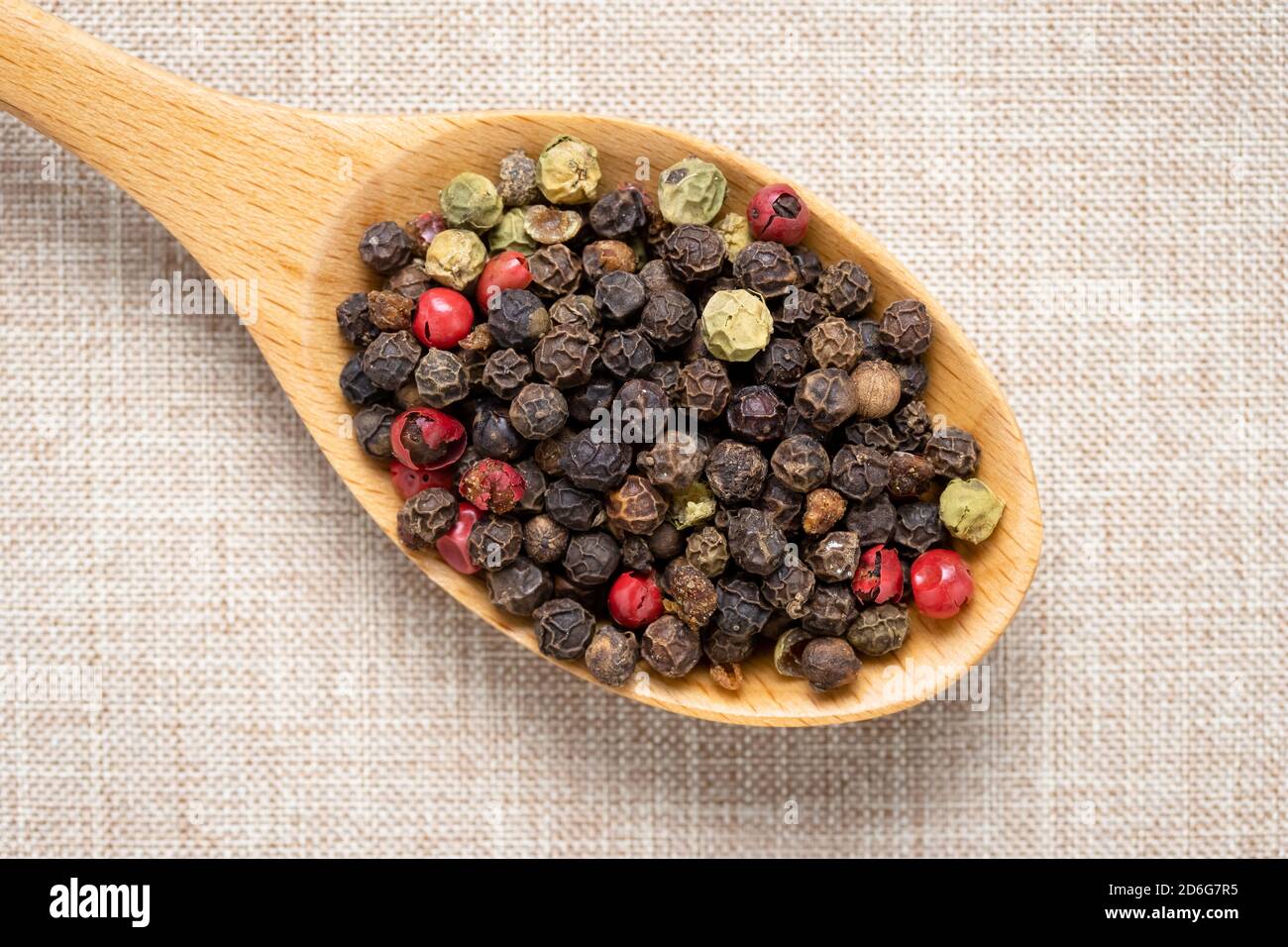 Pepper in a wooden spoon on light brown tablecloth. Pink and black peppercorn close-up. Colorful peas heap. Multicolor hot pepper seeds Stock Photo