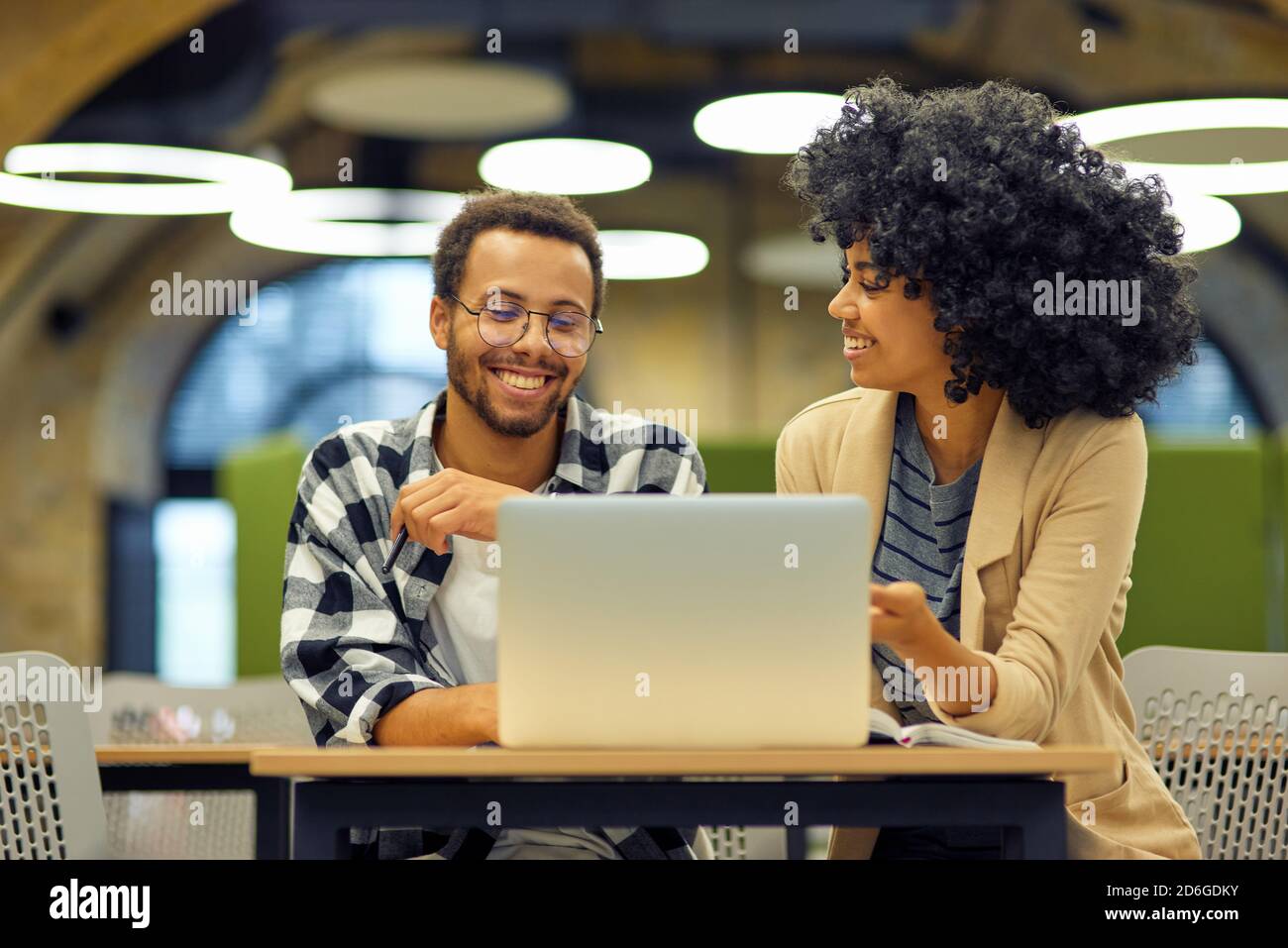 Two young happy multiracial business people sitting at the desk in the modern office, using laptop and discussing project, man and woman working together in coworking space. Office life and business Stock Photo