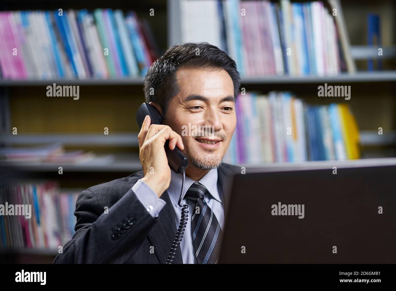 asian corporate executive sitting at desk in office conversing on phone Stock Photo
