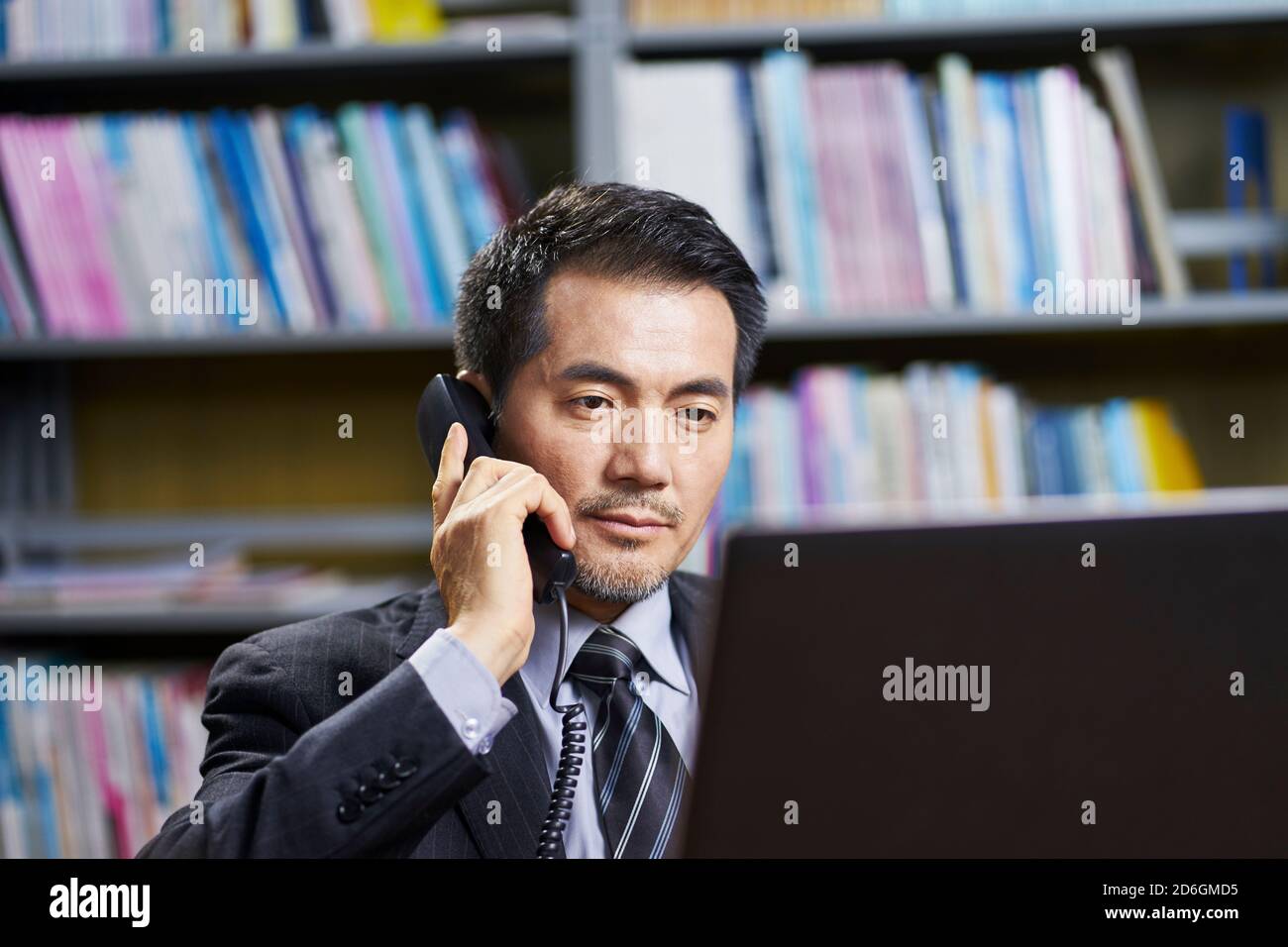 asian corporate executive sitting at desk in office conversing on phone Stock Photo