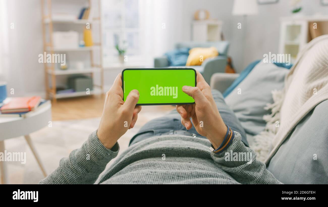 Man at Home Lying on a Couch using Smartphone, Holds it Horizontally in Landscape Mode. Playing Video Games, Watching Videos. Point of View Shot. Stock Photo