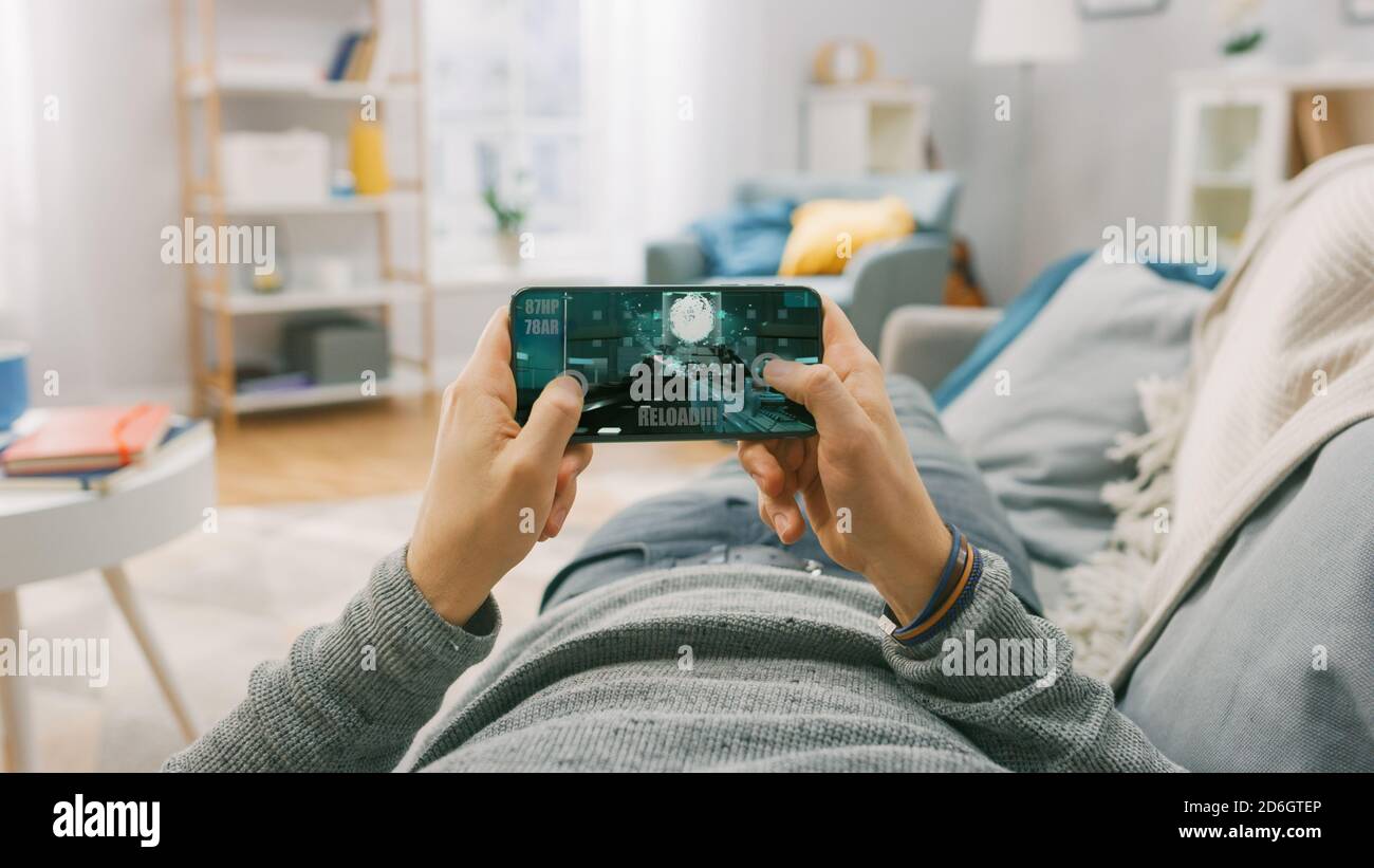 Man at Home Lying on a Couch using Smartphone, Holds it Horizontally in Landscape Mode. He is Playing First Person Shooter Video Game. Point of View Stock Photo