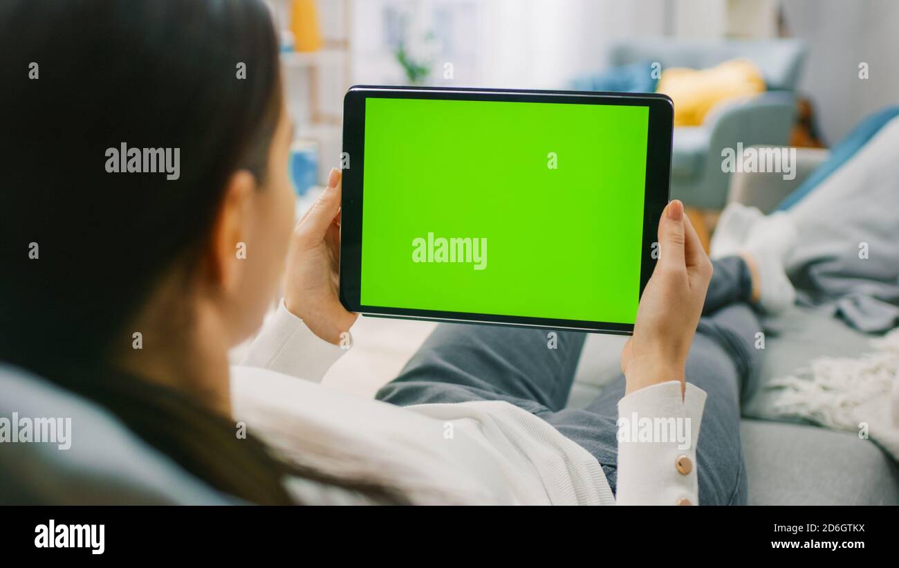 Young Woman at Home Resting on a Couch Using with Green Mock-up Screen Tablet Computer in Horizontal Landscape Mode. Woman Using Gestures with Stock Photo