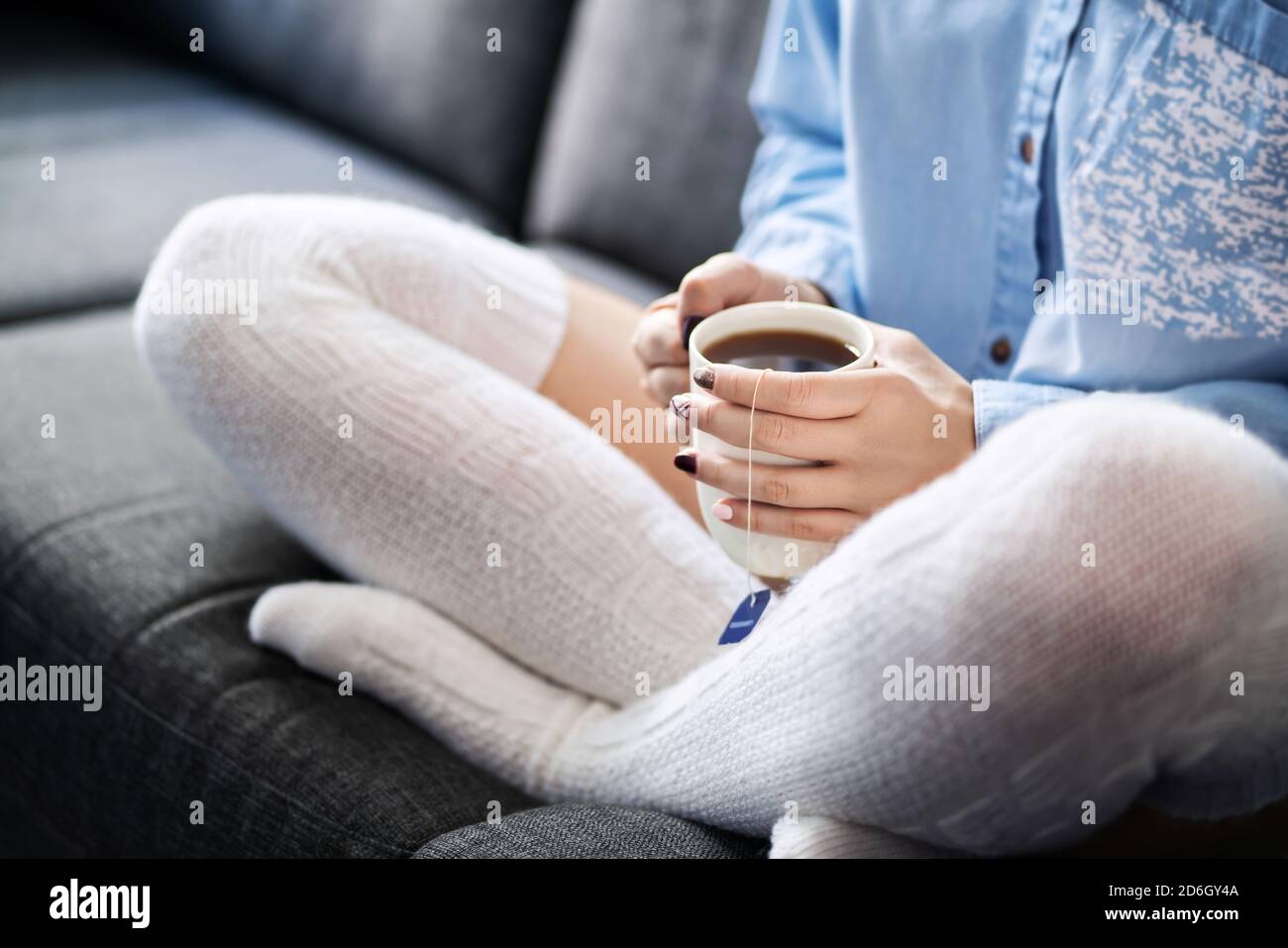Long cozy warm socks and a hot cup of tea in winter. Woman enjoying relaxed lifestyle on home couch. Sick person with flu or fever sitting on sofa. Stock Photo
