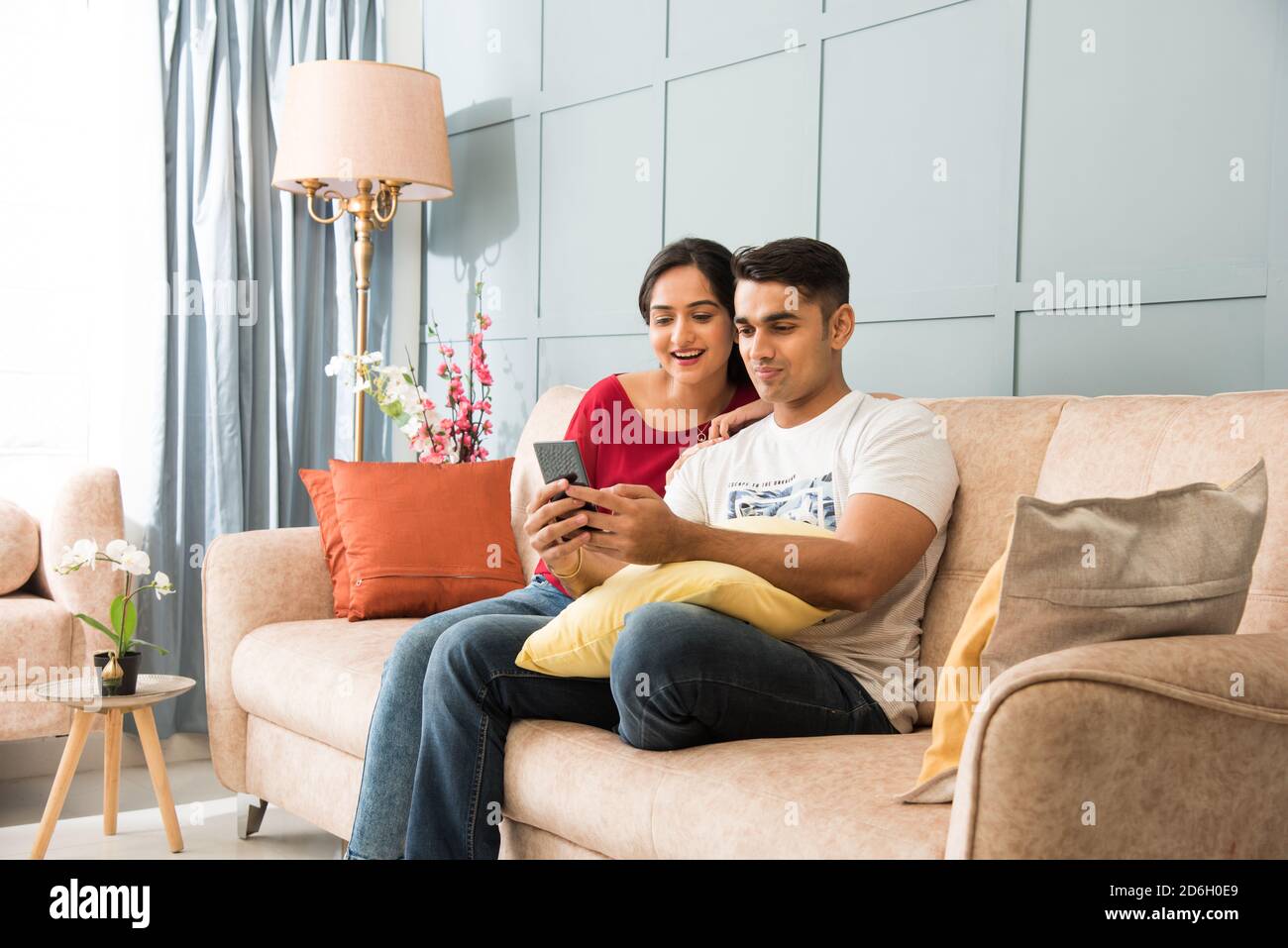 Indian asian young couple using smartphone together while sitting on sofa or couch and modern interior space or home Stock Photo