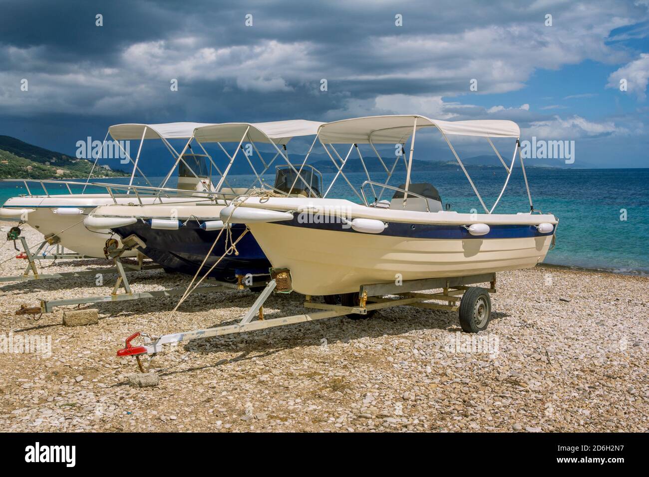 White cruise boats on trailers for transportation on the beach – turquoise sea water, gray clouds on the sky and mountains on the horizon Stock Photo