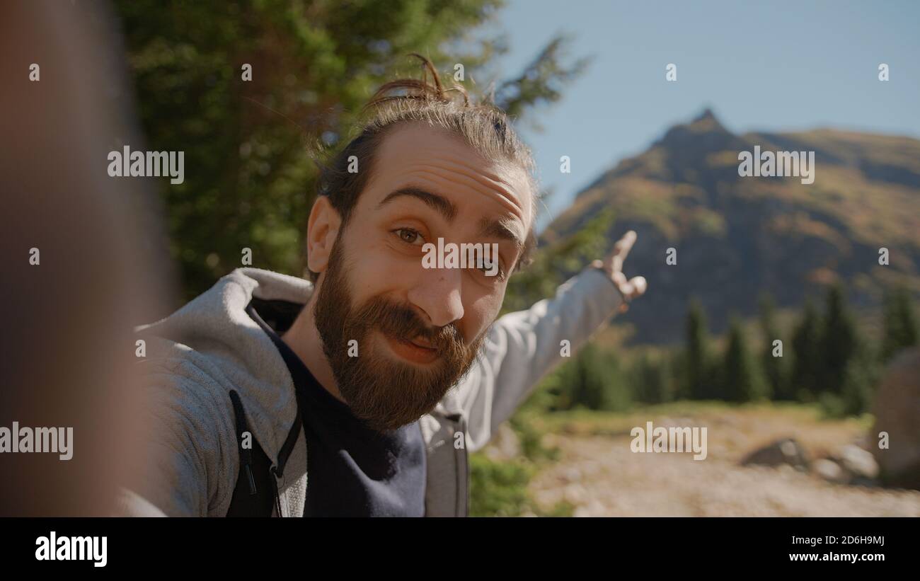 Hippie climber with his backpack behind him, with his handheld camera or phone, salutes the audience or the person he is talking and points to a high Stock Photo
