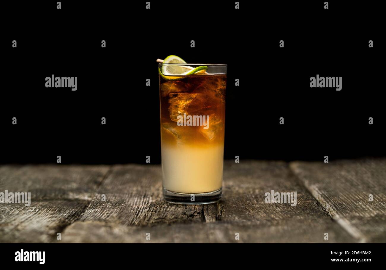 Dark and Stormy cocktail in high ball glass with ice and a lime garnish on an old wooden table with a black background Stock Photo