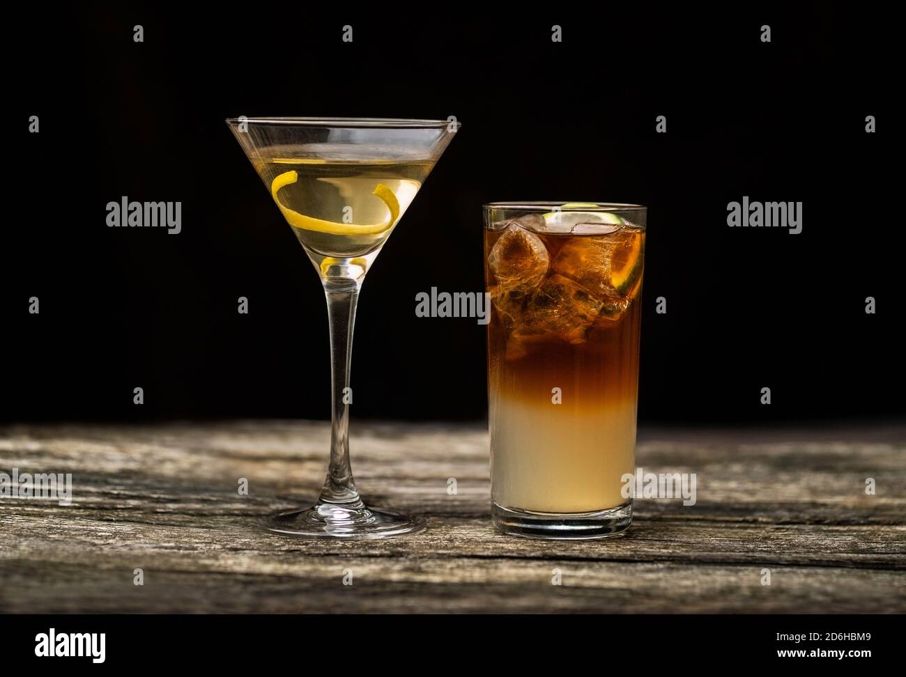 Dark and Stormy cocktail in high ball glass with ice and a lemon garnish with a Vodka Martini on an old wooden table with a black background Stock Photo