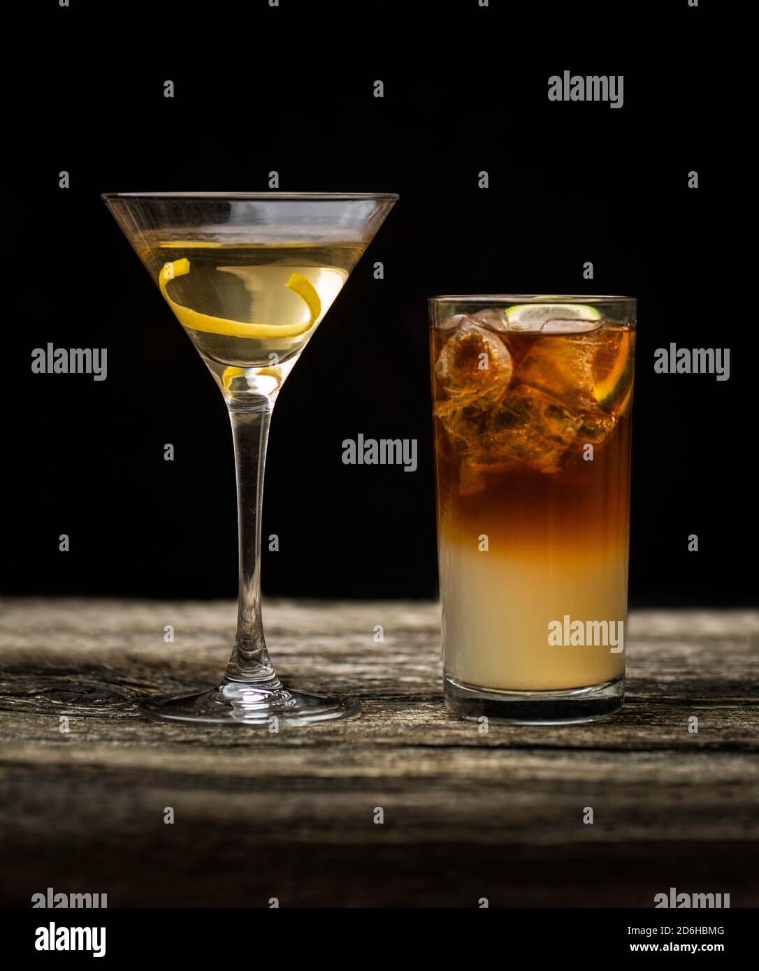 Dark and Stormy cocktail in high ball glass with ice and a lemon garnish with a Vodka Martini on an old wooden table with a black background Stock Photo