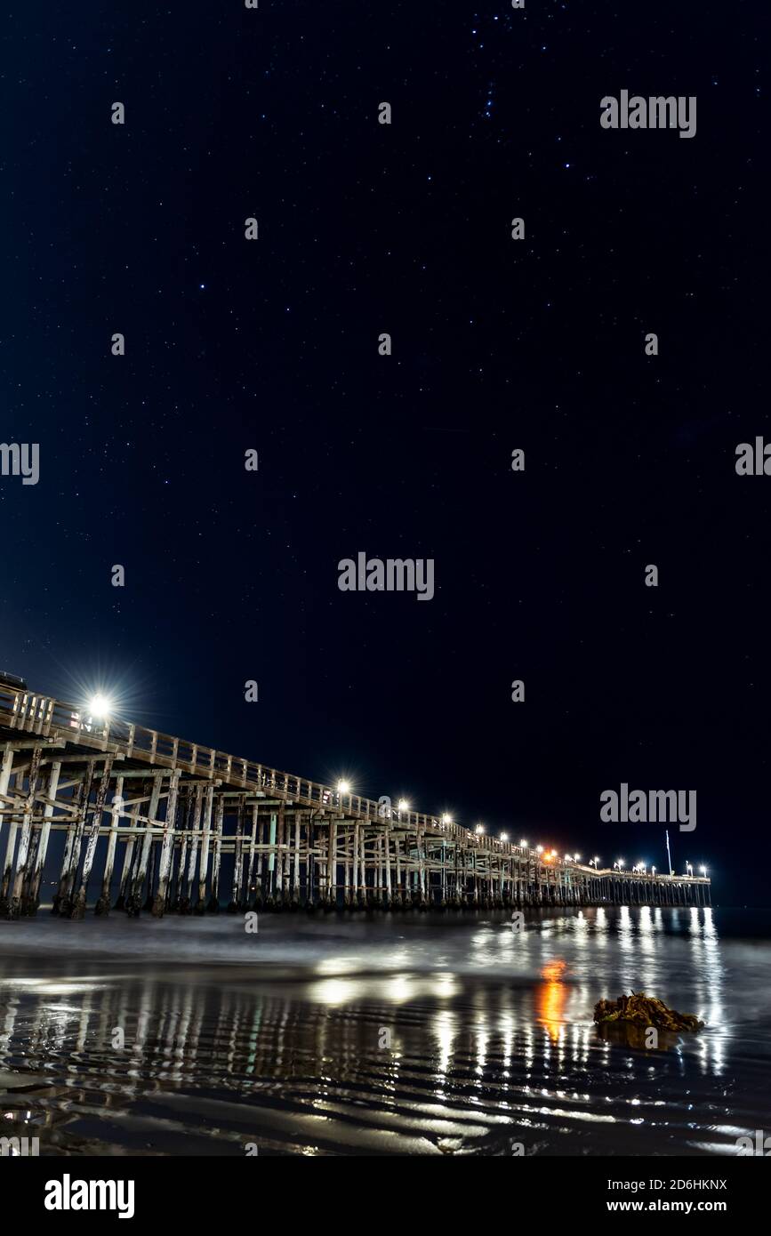 Ripple textures in the wet sand as lamps reflect in the ocean water under the Ventura Pier with constellation stars in sky above. Stock Photo