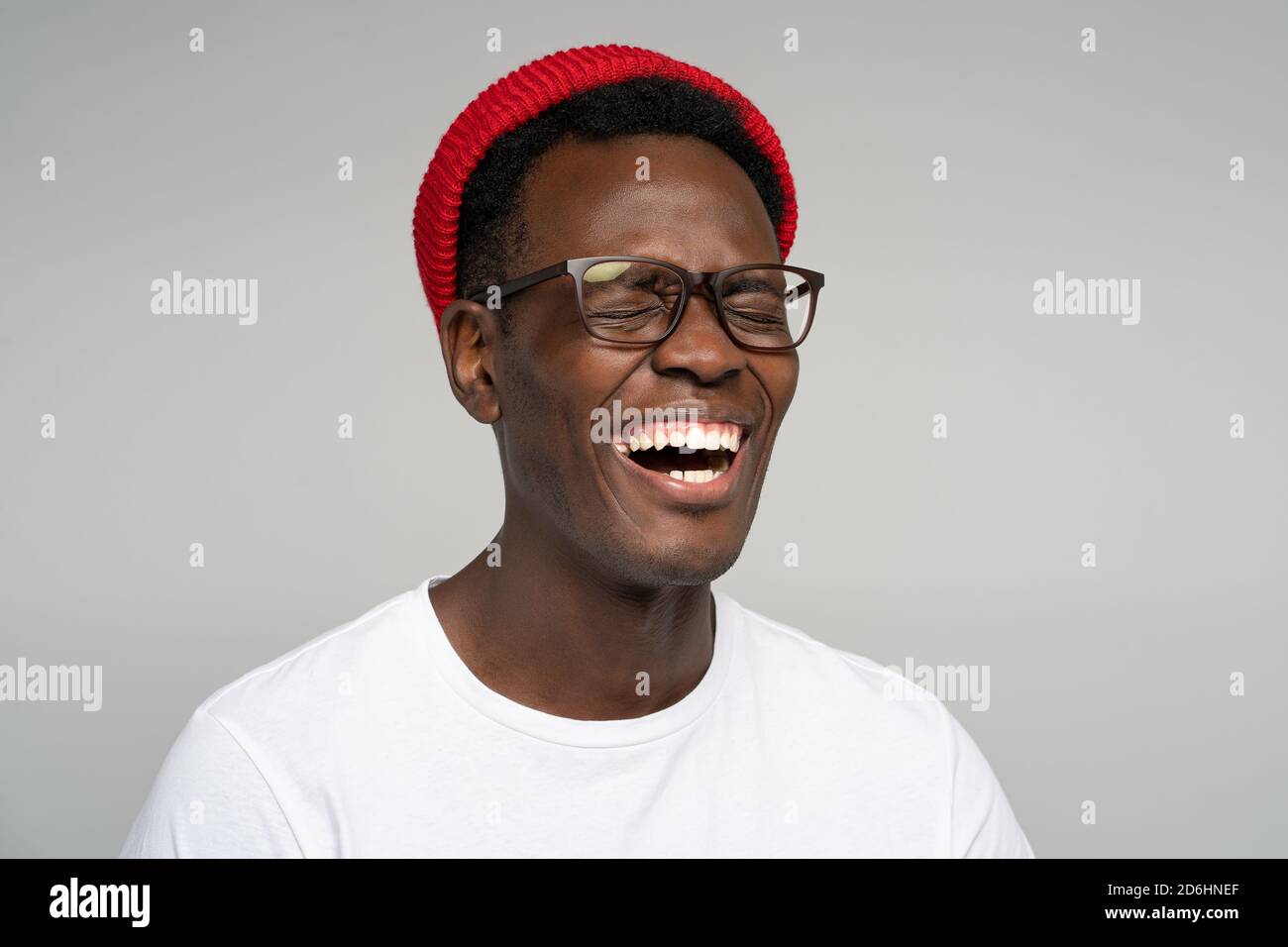 Laughing positive young Afro American man wear red hat in good mood, poses at camera with closed eyes. Overjoyed Black male in spectacles shows her br Stock Photo
