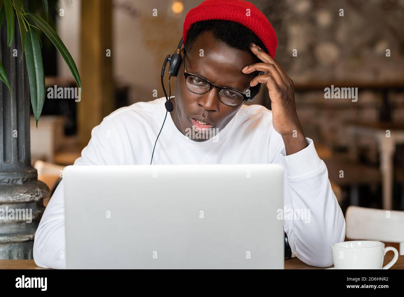 Thoughtful millennial Black man in headphones wear red hat, watching educational webinar on laptop, remotely online work in cafe. Afro-American male l Stock Photo