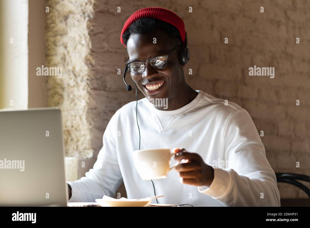 Happy millennial Black man in headphones wear red hat, enjoying watching educational webinar on laptop, remotely online work in cafe, holding cup of c Stock Photo