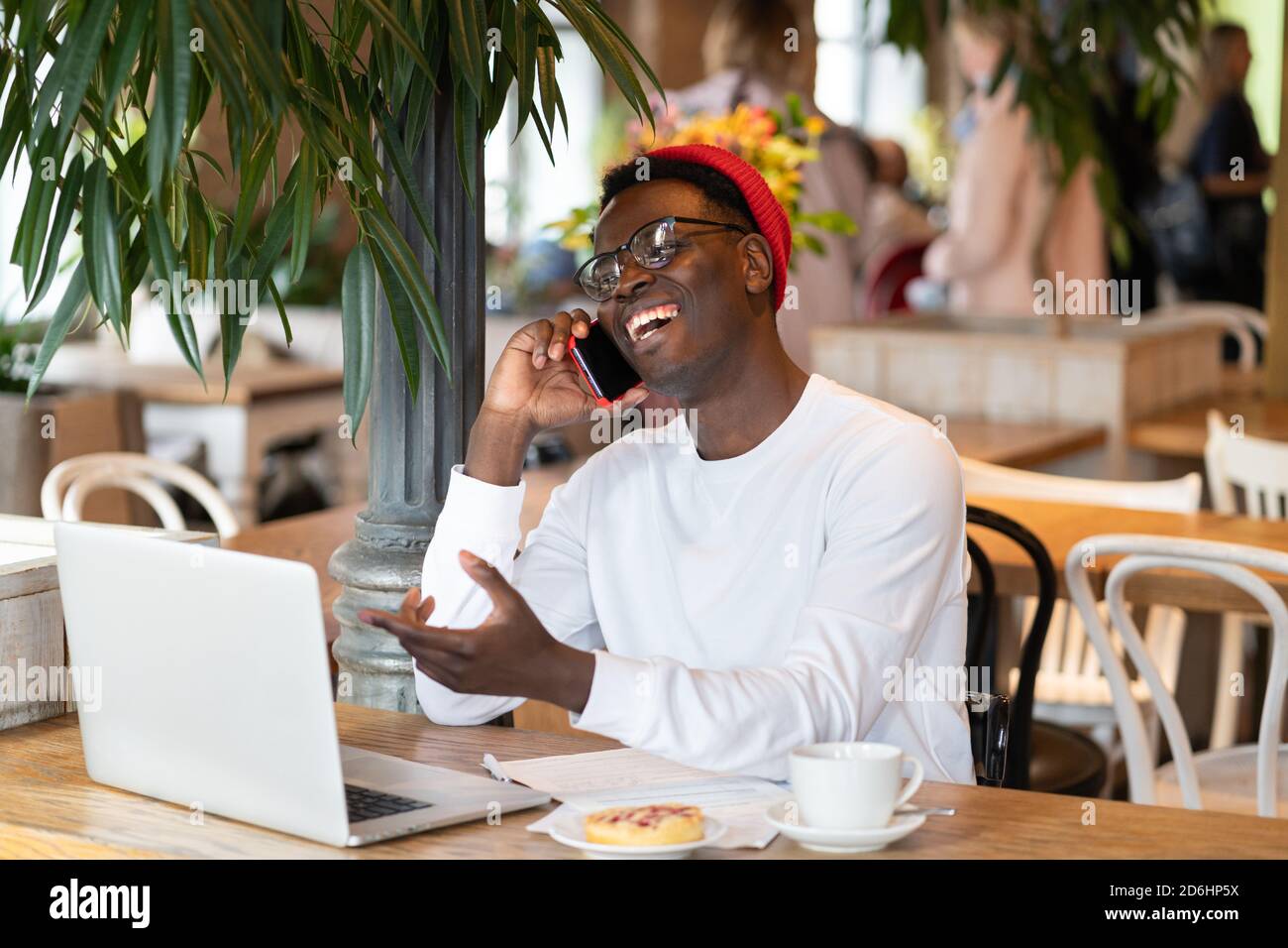 Happy millennial Afro-American man wear red hat laughing, speaking with friend on mobile phone, enjoying watching educational webinar on laptop, remot Stock Photo